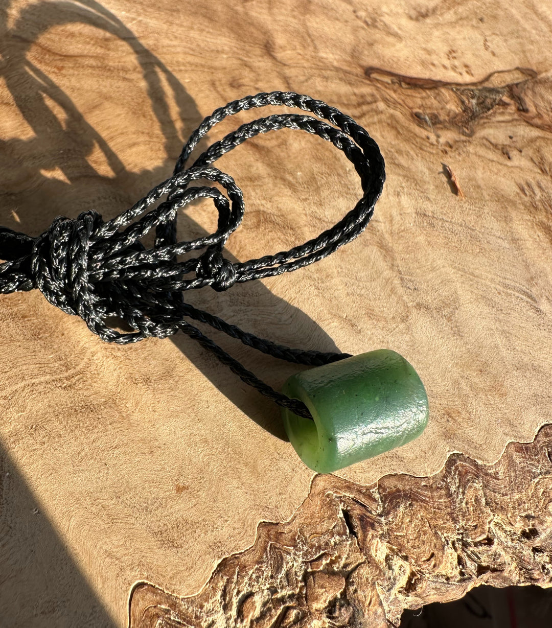 Close-up of a textured green jade pendant threaded on a black braided cord with a knot on a wooden surface