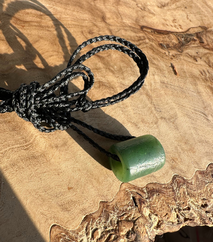 Close-up of a textured green jade pendant threaded on a black braided cord with a knot on a wooden surface