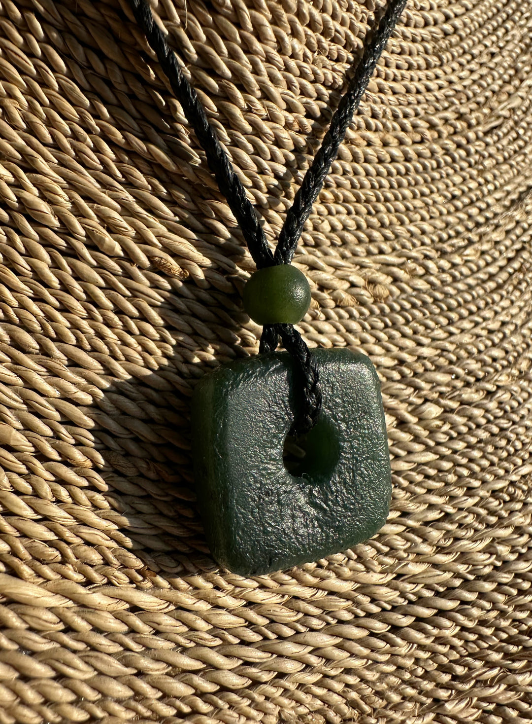 Canadian Jade textured square pendant with a central hole on a black braided cord against a woven straw background.