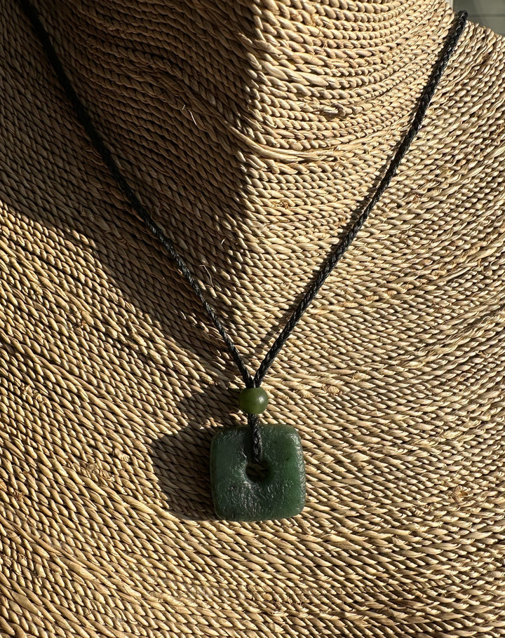 Canadian Jade textured square pendant with bead on a black braided cord displayed on a woven straw backdrop.