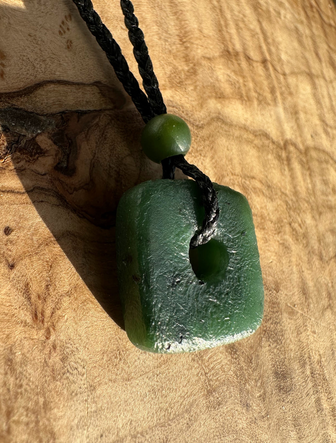 Close up of Canadian Jade textured square pendant with a hole and bead on a black cord resting on a wood surface.