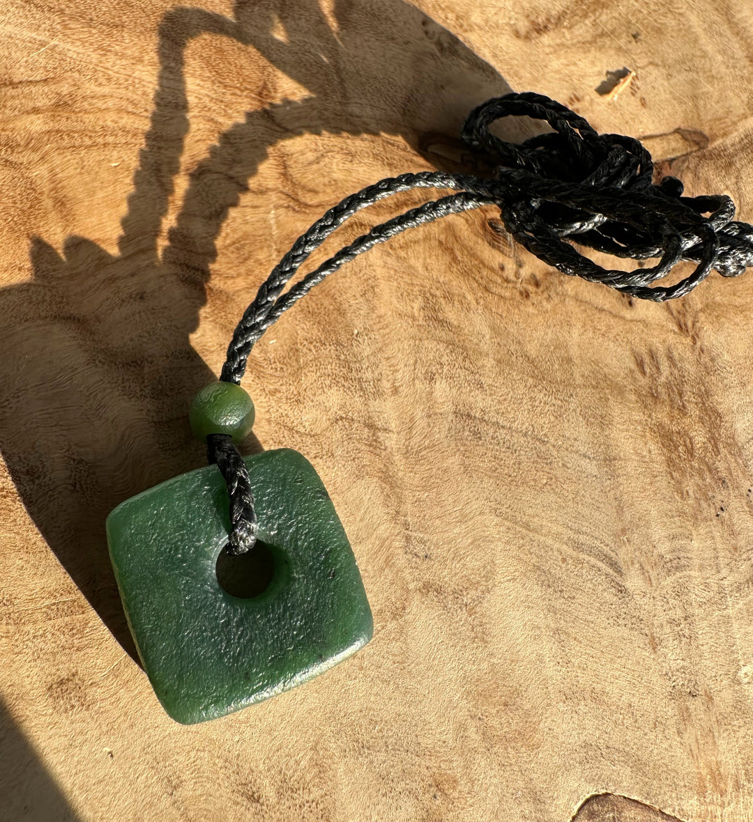 Green jade square pendant with a central hole on a braided black cord and a small green bead