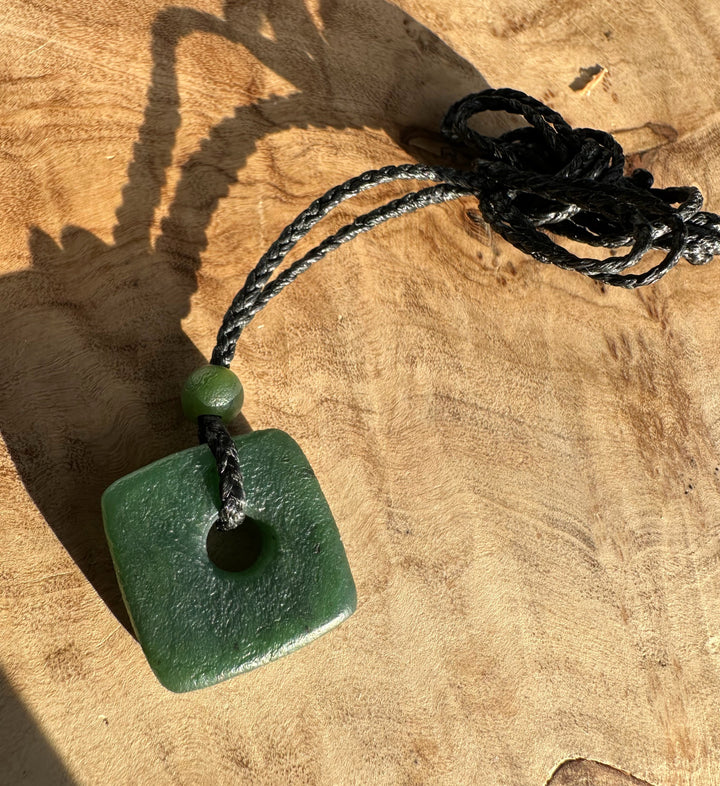 Green jade square pendant with a central hole on a braided black cord and a small green bead