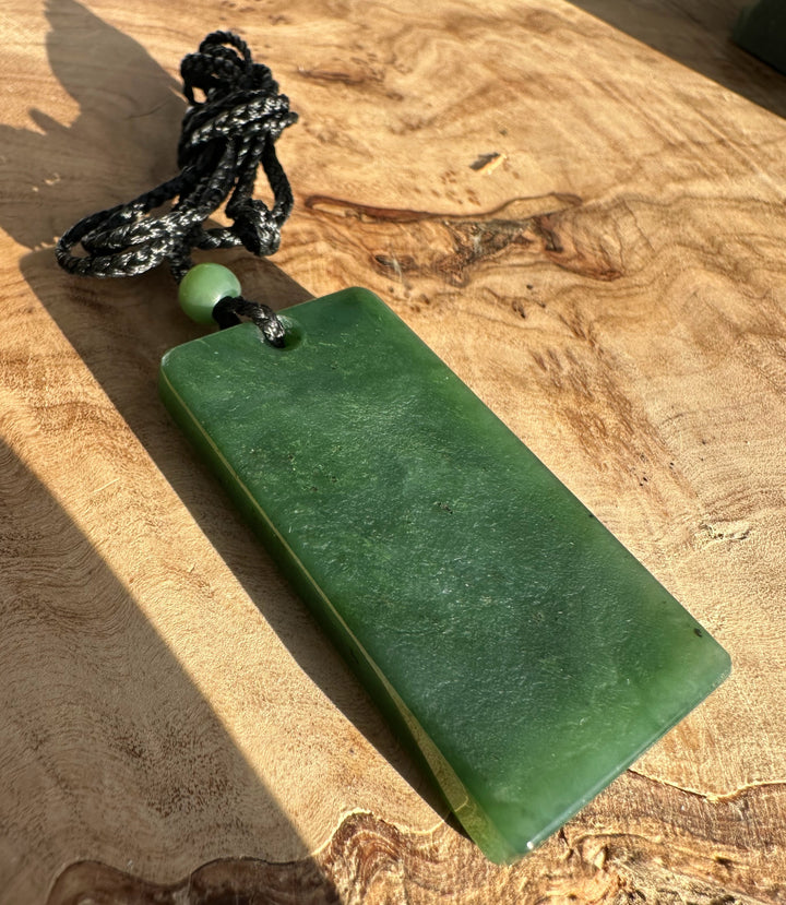 Canadian Jade Textured Rectangle Pendant 50MM on a black braided cord with a green bead resting on wood