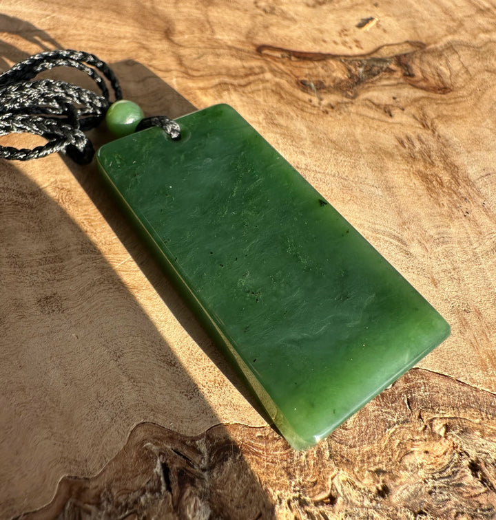 Canadian Jade Textured Rectangle Pendant 50MM with black cord and bead on a wooden surface