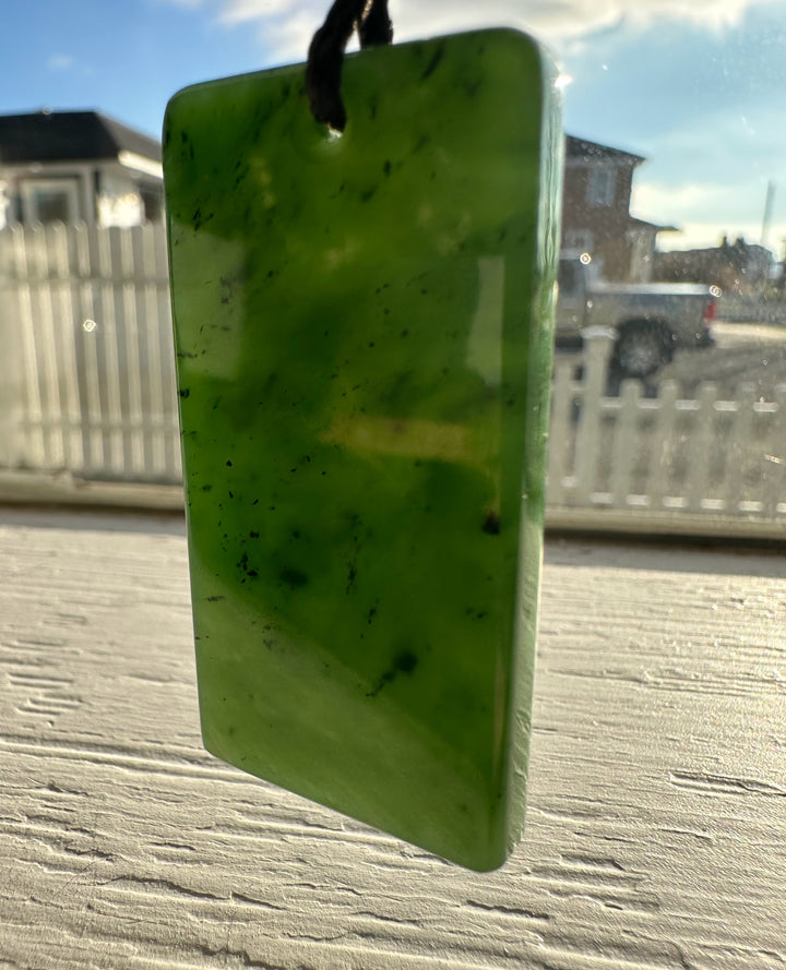 Canadian Jade Textured Rectangle Pendant 50MM suspended from a cord against a sunny window backdrop