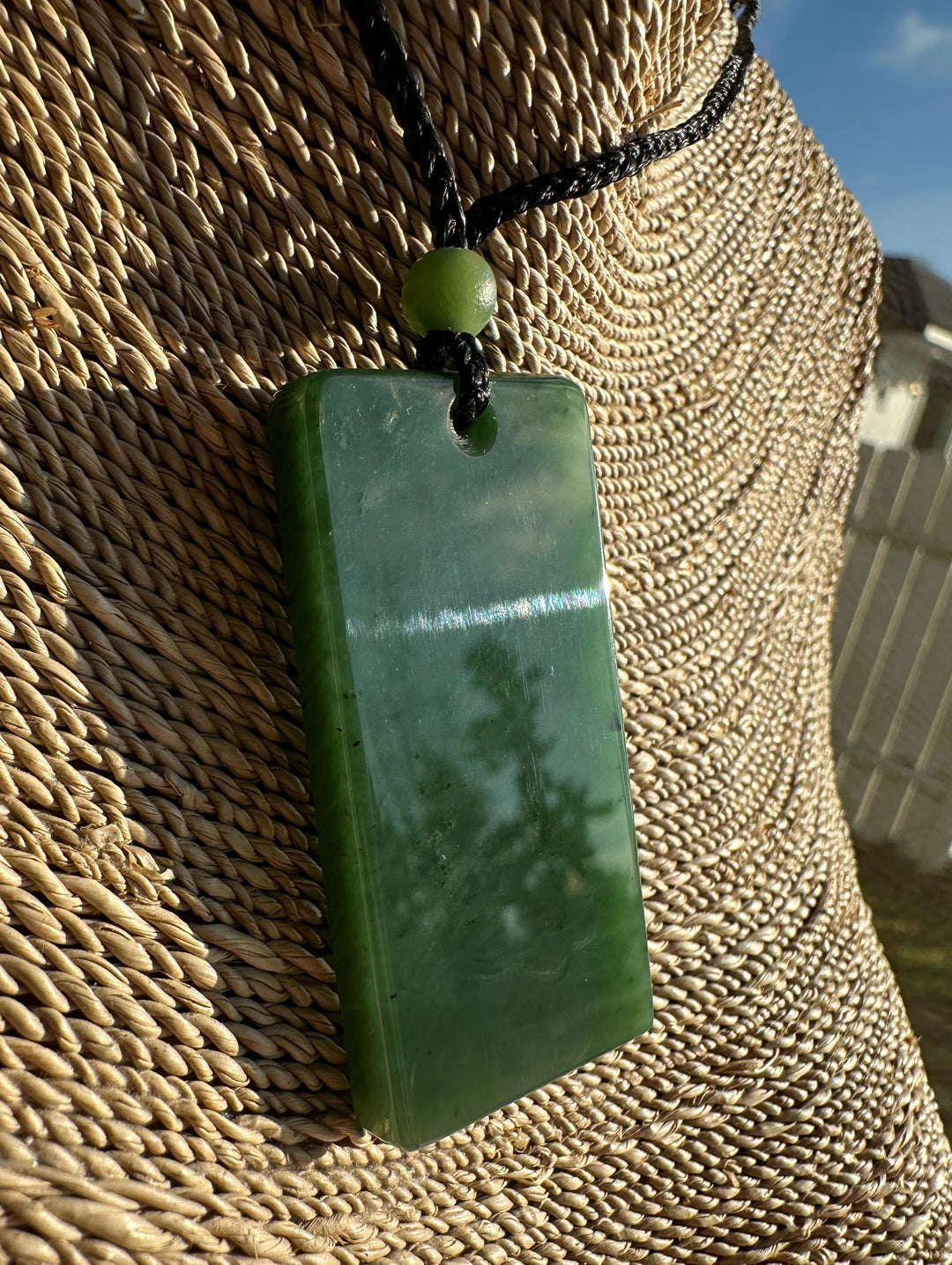 Green rectangular jade pendant on a black cord with a green bead against a woven straw background.