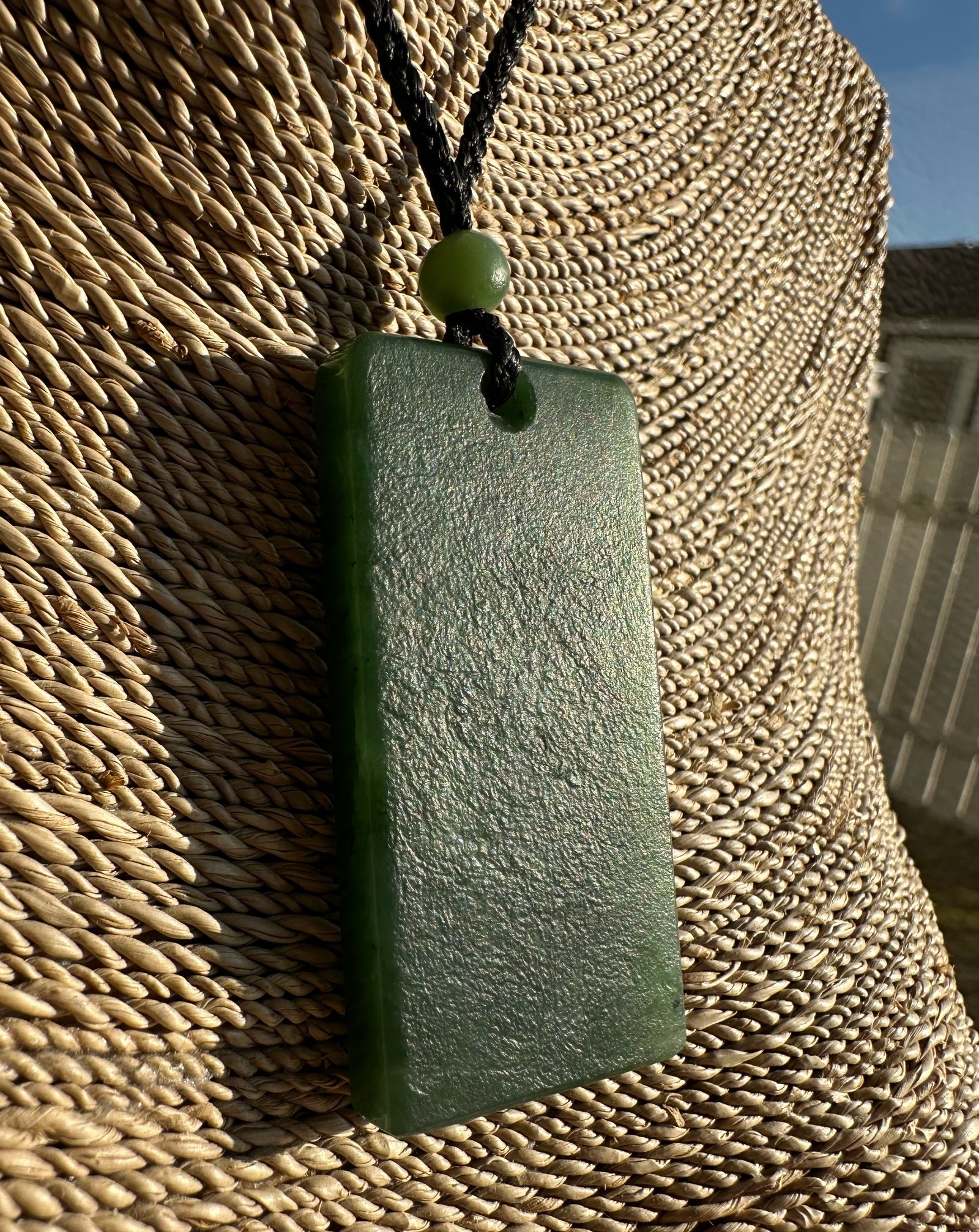 Rectangular jade pendant on black cord with green bead against woven straw background