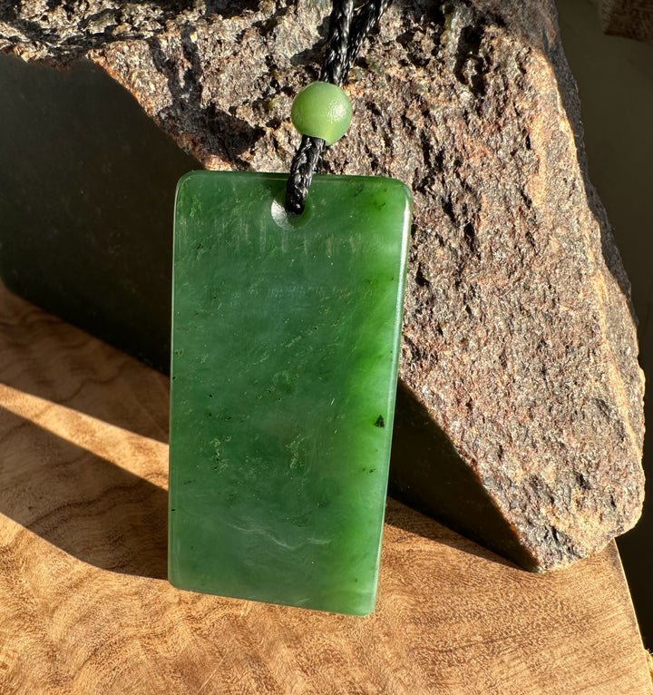 Canadian Jade rectangle pendant with a hole at the top on a black cord and a green bead against a rough rock backdrop.