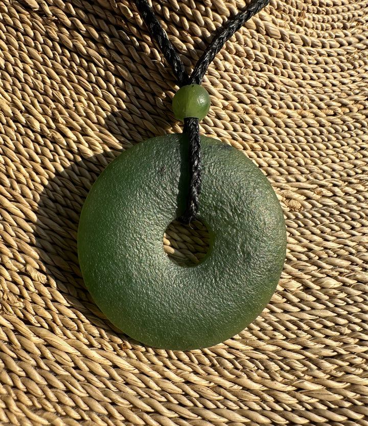 Close up of textured green jade donut pendant on a black cord with bead against woven background