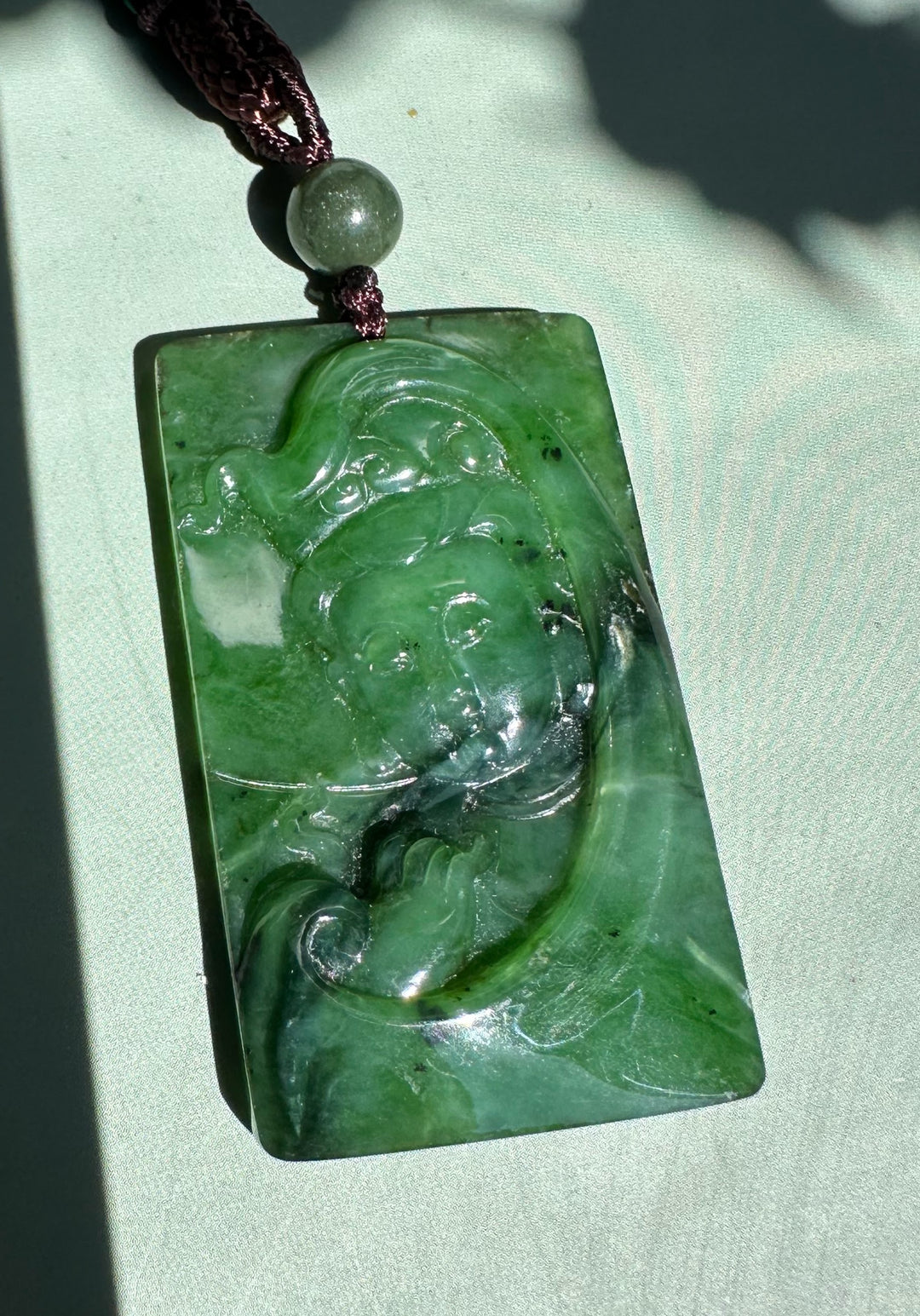 Canadian Jade Guanyin pendant carved from emerald green jade suspended by a brown cord with a round bead on pale green fabric