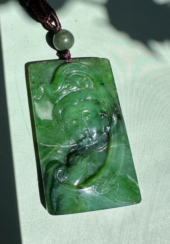 Canadian Jade Guanyin pendant carved from emerald green jade suspended by a brown cord with a round bead on pale green fabric