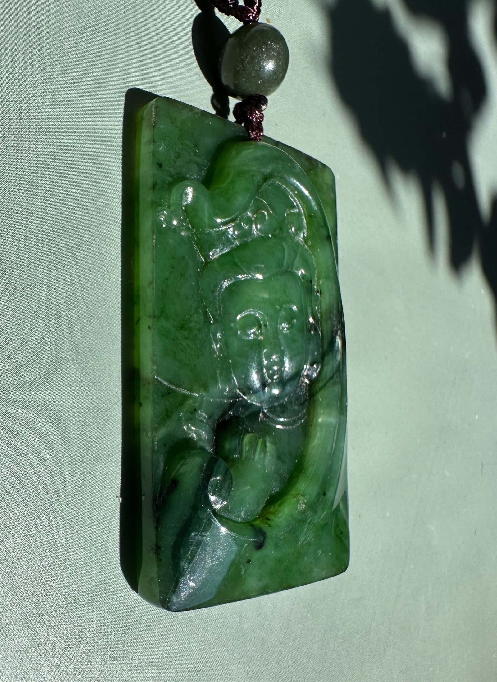 Rectangular green jade pendant carved with Guanyin and suspended on a dark brown cord with a bead.