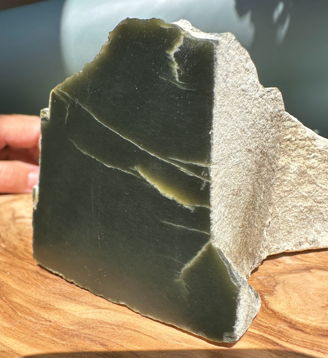 Hand holds a dark olive jade slab with a pale beige crust along the top