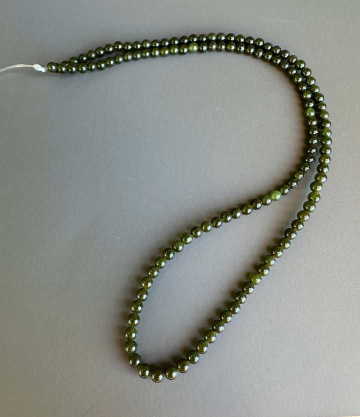 Dark green 3mm nephrite jade beads on an unstrung strand coiled against a gray backdrop.