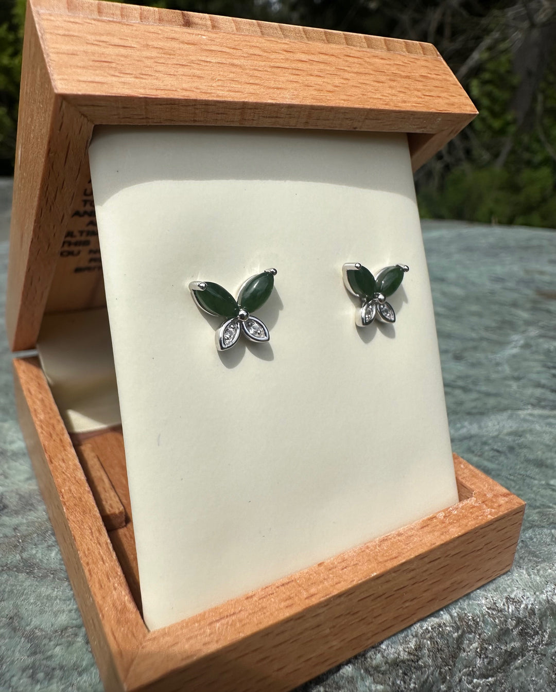 Angled view of silver butterfly studs with green enamel wings and crystal accents mounted on a cream display inside an open wooden box