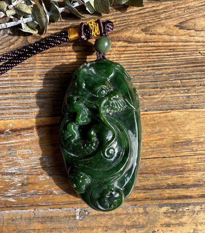 Green jade dragon pocket pendant with swirling carved dragon motifs, hung on a dark brown braided cord with a green bead.