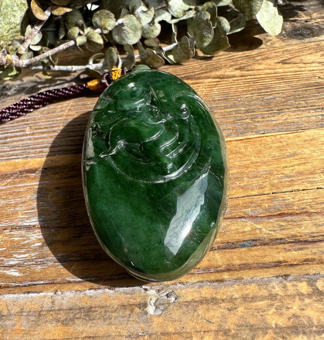 Deep green jade dragon pocket pendant shown from above, polished surface with carved swirls on a wooden backdrop and cord.