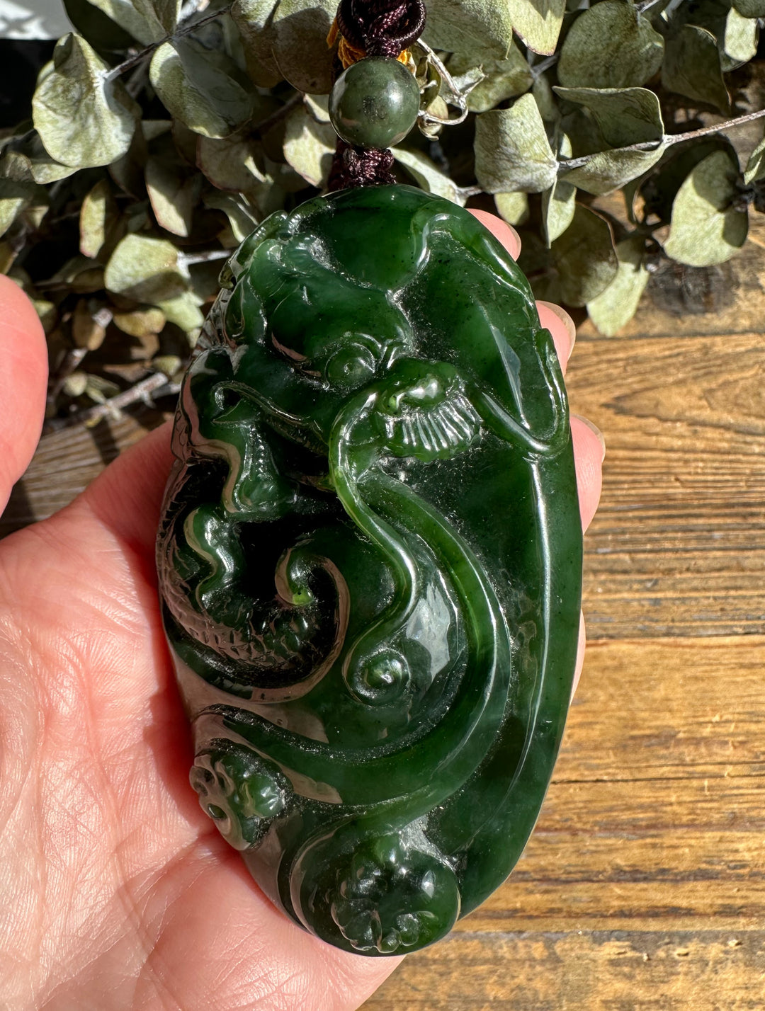 Handheld view of green jade dragon pocket pendant with braided cord and bead, showcasing intricate carving.