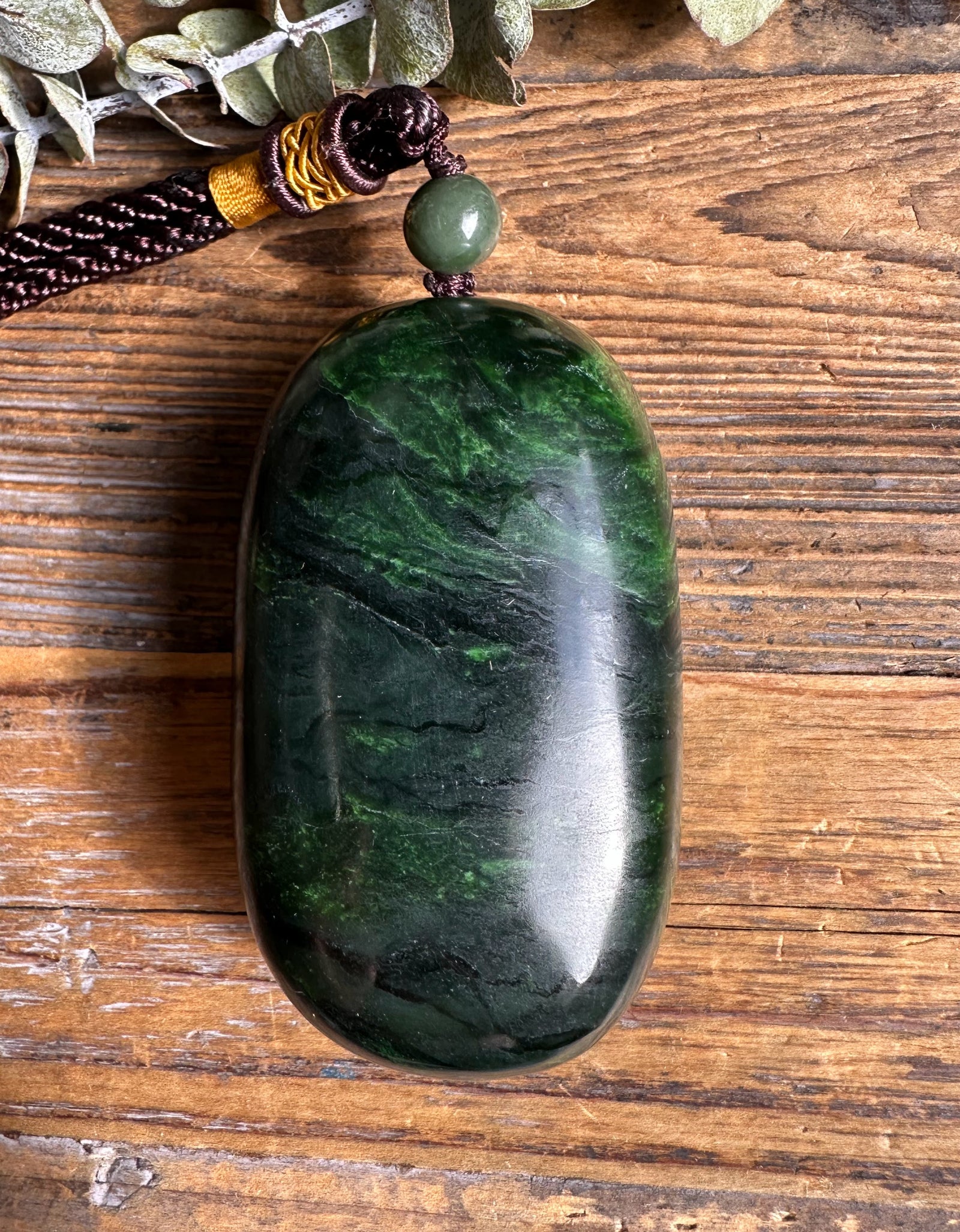 Back view of a smooth dark green oval jade pendant on a brown braided cord with a bead at the top