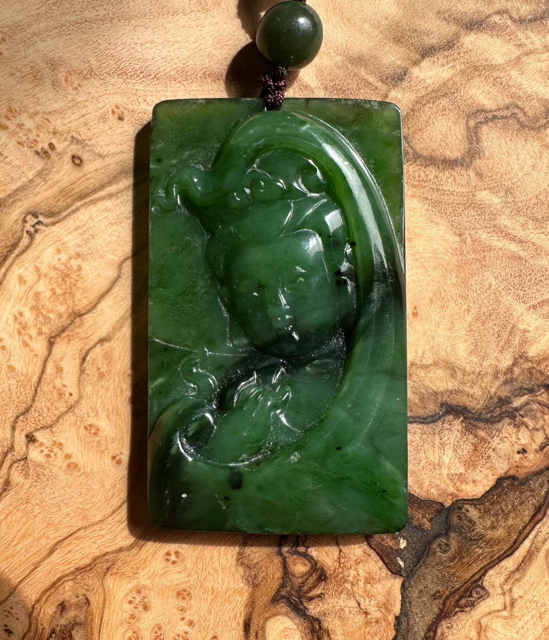 Canadian Jade Guanyin pendant carved in green jade, rectangular slab with a brown cord and bead above on a wood surface