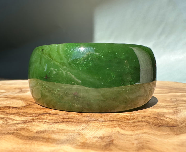 Wide Cassiar jade bangle in two tone green with a lighter base on a wooden surface