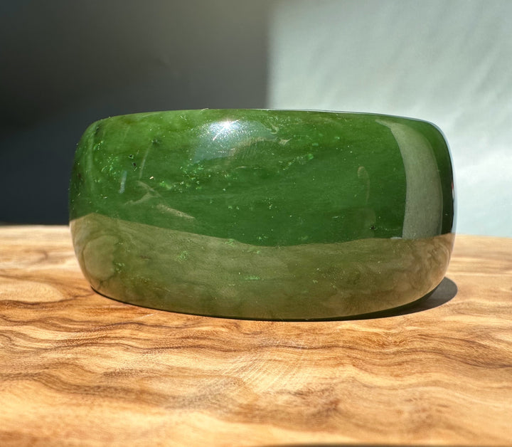Side view of Wide Cassiar Jade Bangle with green exterior on a wooden surface