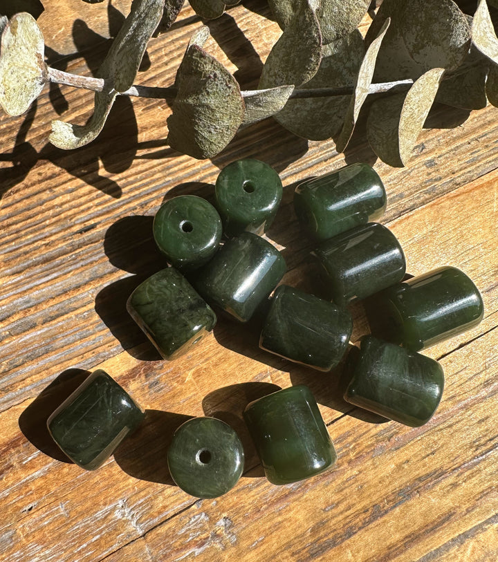 Group of dark green jade barrel beads 13x15mm with holes on a wooden surface among dried leaves