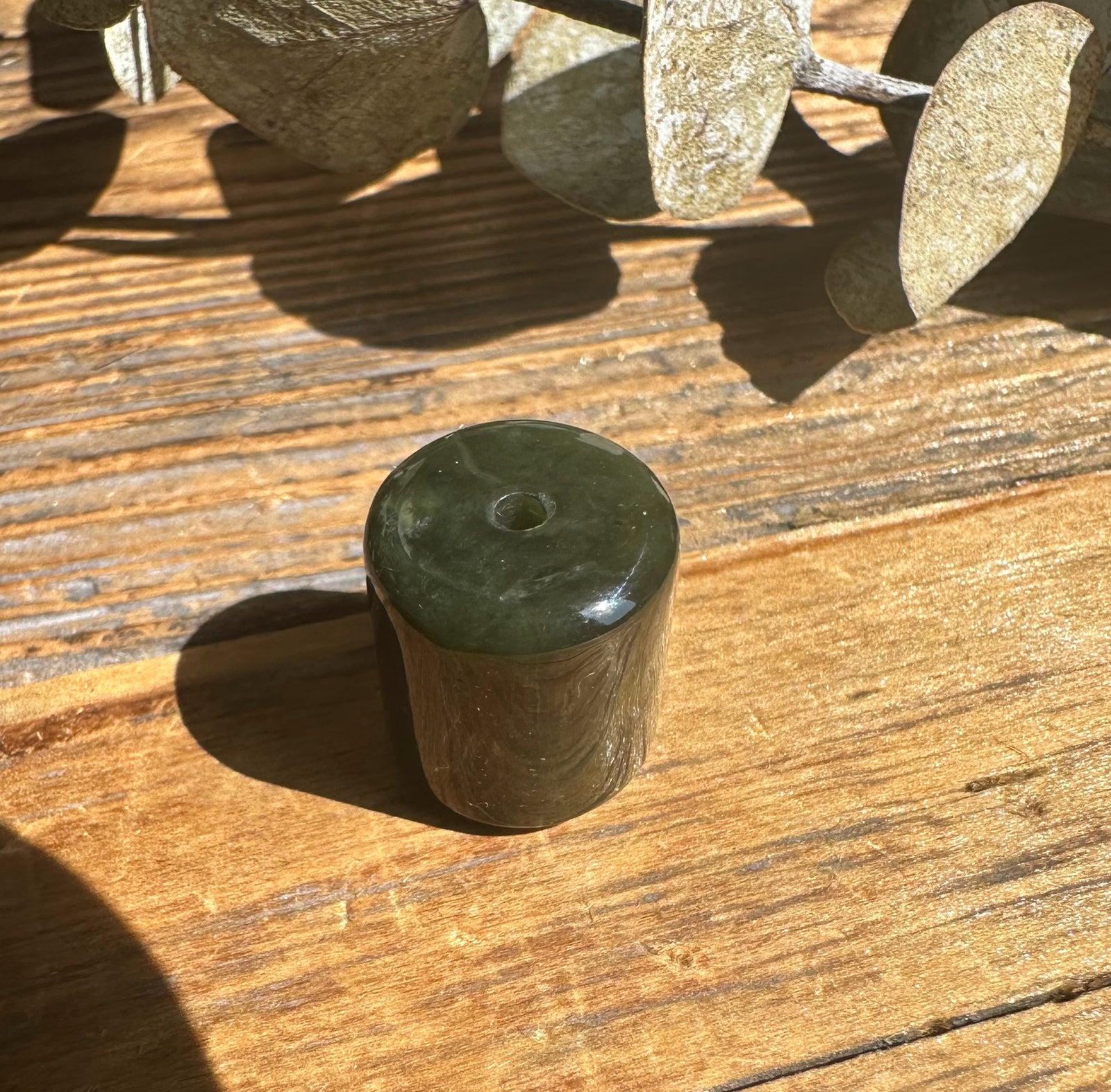 Close up of a single dark green jade barrel bead with a drilled hole on a sunlit wooden surface
