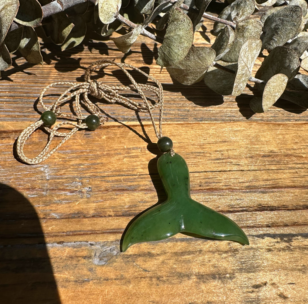 Jade whale tail pendant with a beige braided cord and green beads on a wooden surface