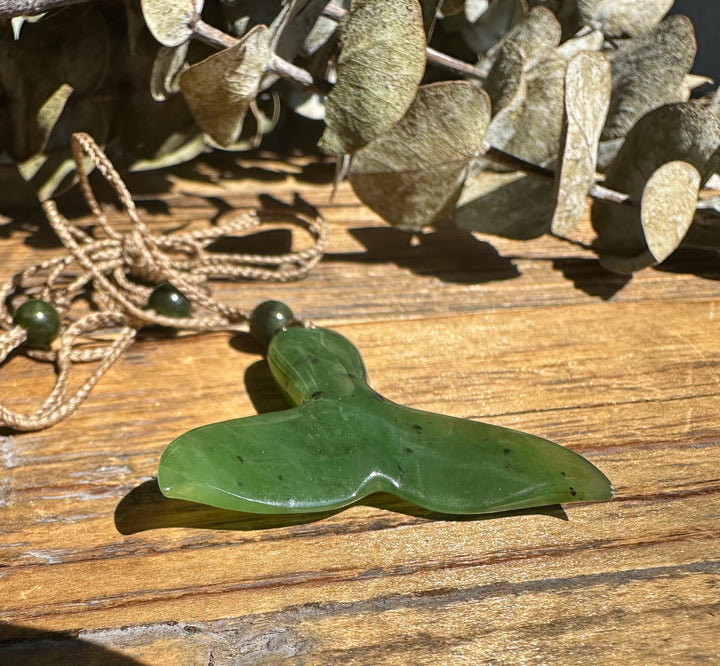 Green jade whale tail pendant with cord and bead resting on a wooden surface.