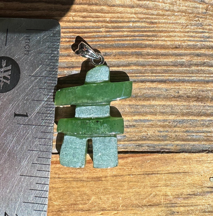 Green jade Inukshuk pendant with a silver bail on a cord beside a ruler for scale.
