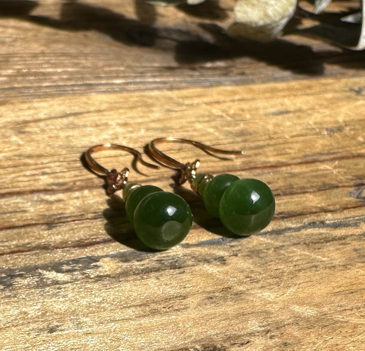 Two green jade double-gourd beads with copper findings resting on a wooden surface.