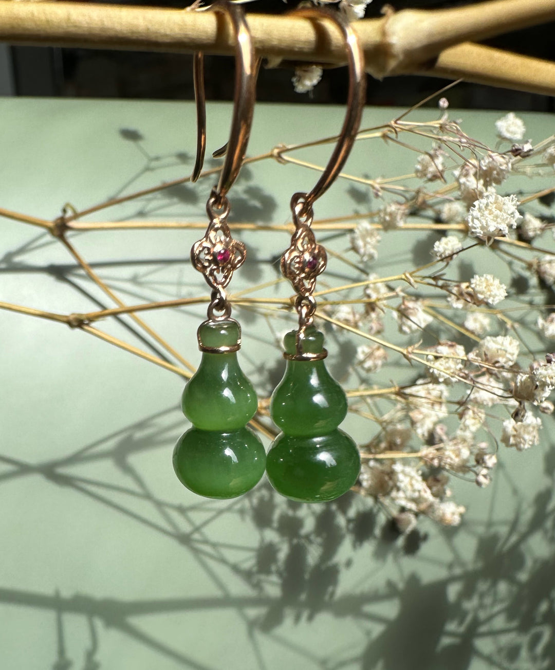 Pair of green jade double-gourd drop earrings with rose gold hooks against a rustic background.