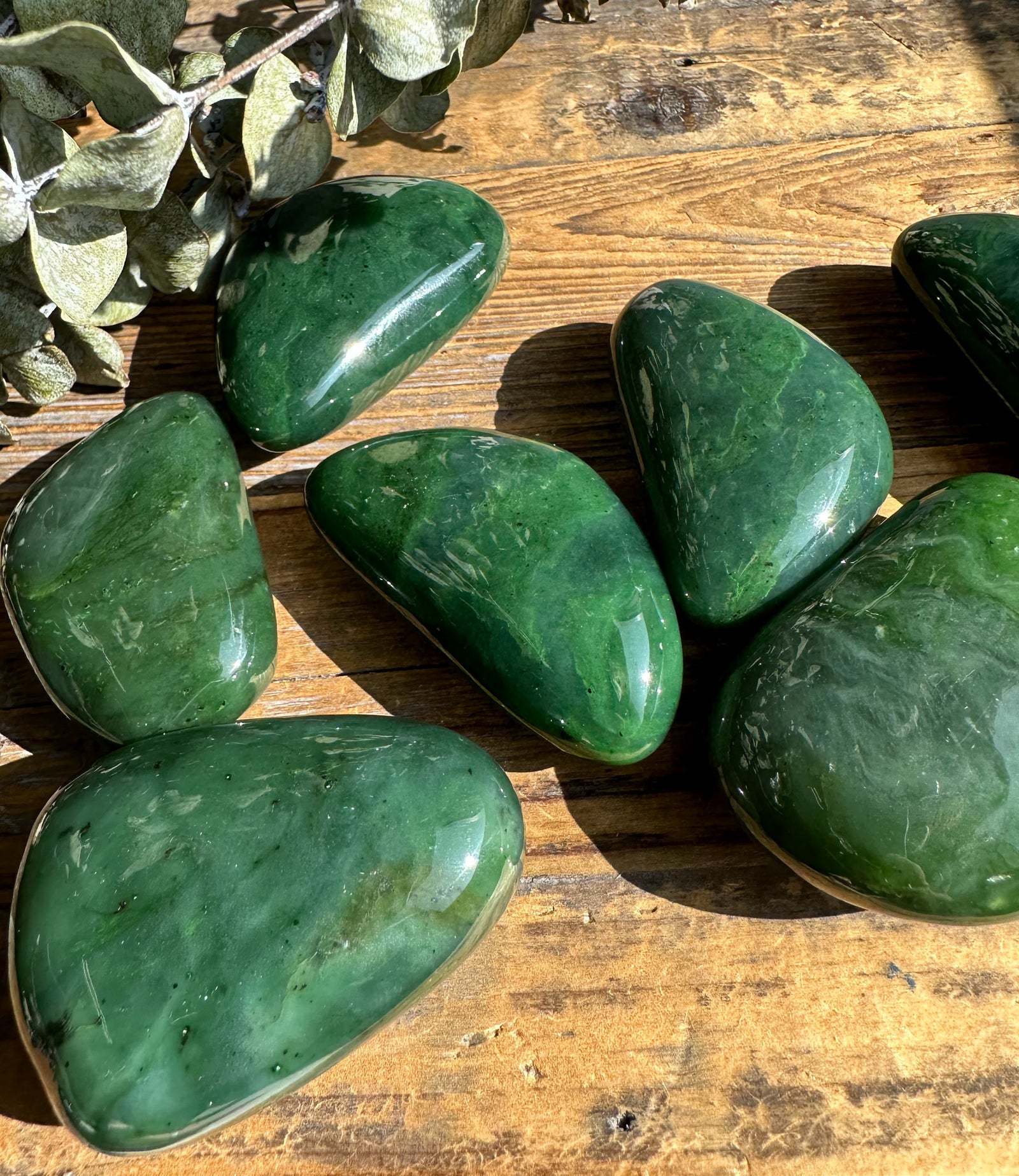 Polished jade specimens in varying shapes resting on a wooden surface beside dried eucalyptus leaves.