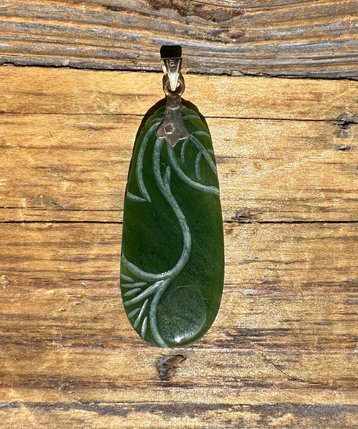 Close up of oval green jade pendant featuring a carved vine motif and gold bail on wood