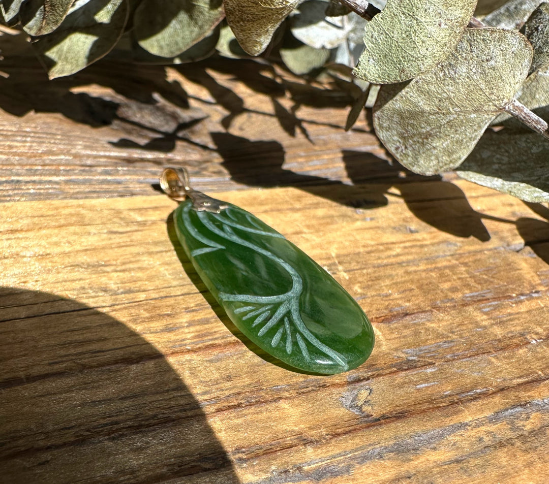 Green jade pendant carved with a flowing white leaf and vine pattern, smooth oval shape with a gold bail on wood