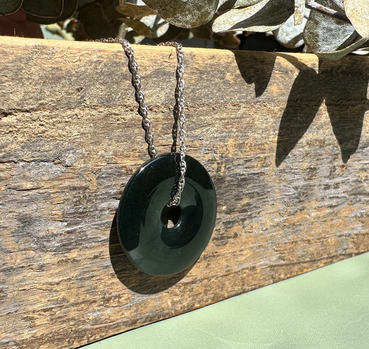 Dark green Australian Jade donut pendant on a silver chain draped over a weathered wooden surface.