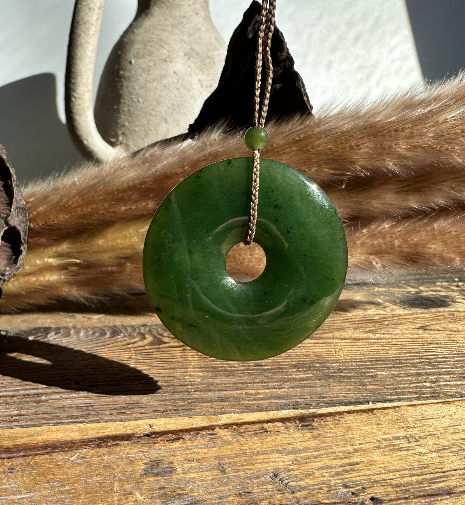 Green jade donut pendant threaded on a brown cord through the center, hanging against a wooden surface and dried grasses.