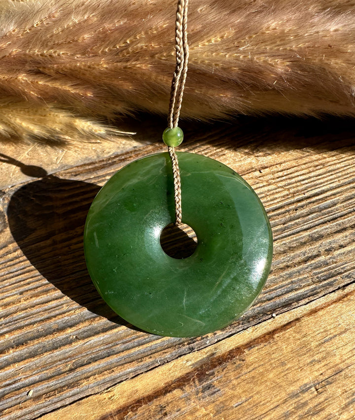 Close up of Canadian Jade PI Donut 45mm pendant suspended on a braided cord with a small green bead, set on a rustic wooden surface.