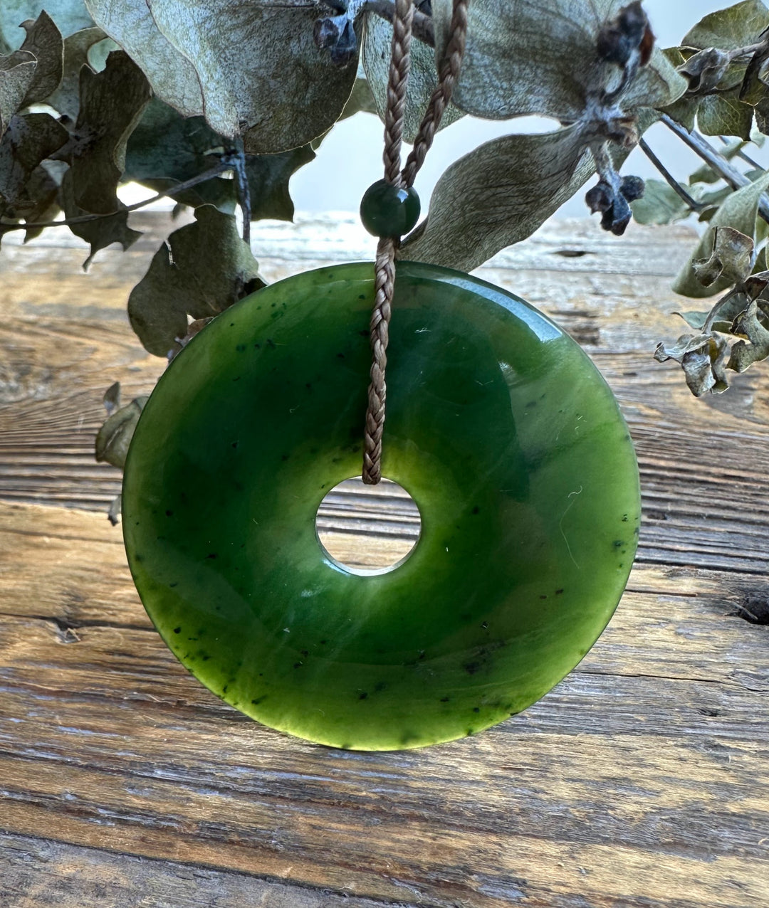 Canadian Jade PI Donut 45mm pendant on a natural braided cord with a green bead, displayed on weathered wood with dried foliage.
