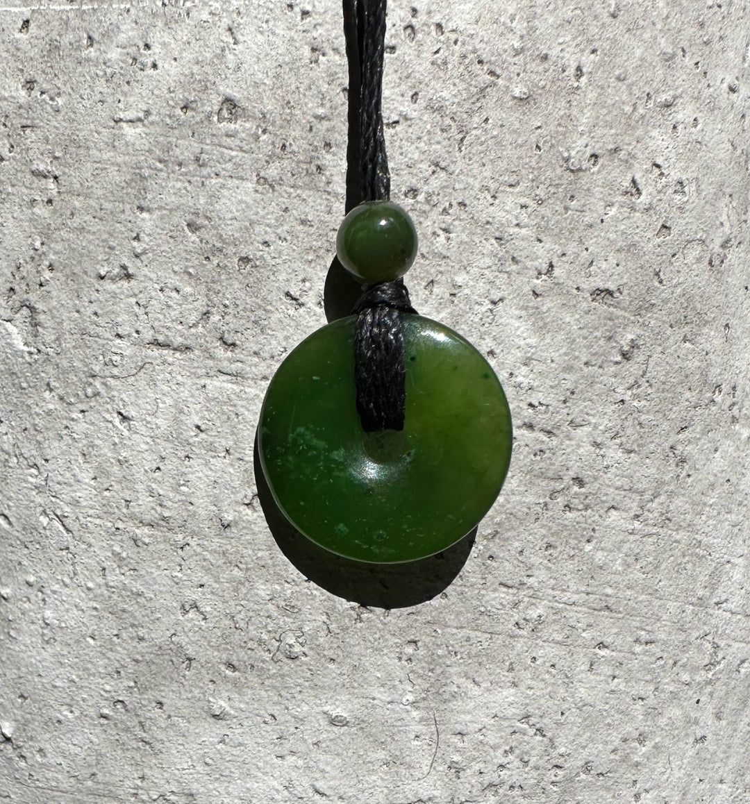 Jade donut pendant with a small bead on a black cord hanging against a textured stone wall.