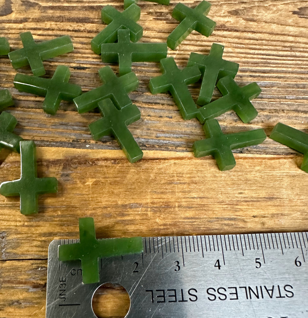Close-up of drilled green jade cross charms, 20mm, scattered near a metal ruler for scale
