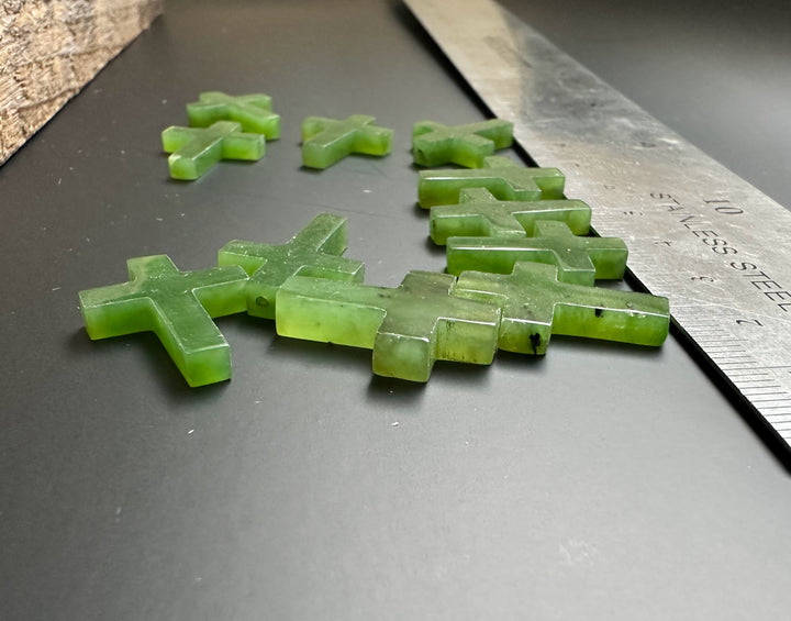 Group of drilled green jade cross charms, 20mm, on a dark surface beside a ruler