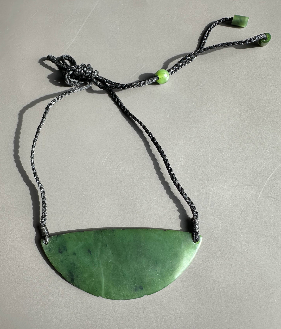 Green jade crescent pendant on knotted black cord with green beads shown on a gray surface