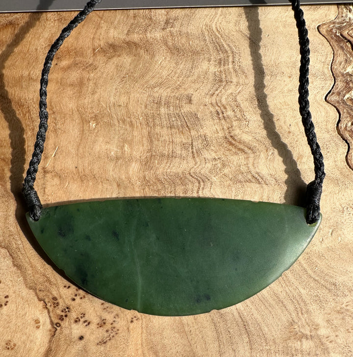 Green jade crescent pendant with a black braided cord on a wooden surface.