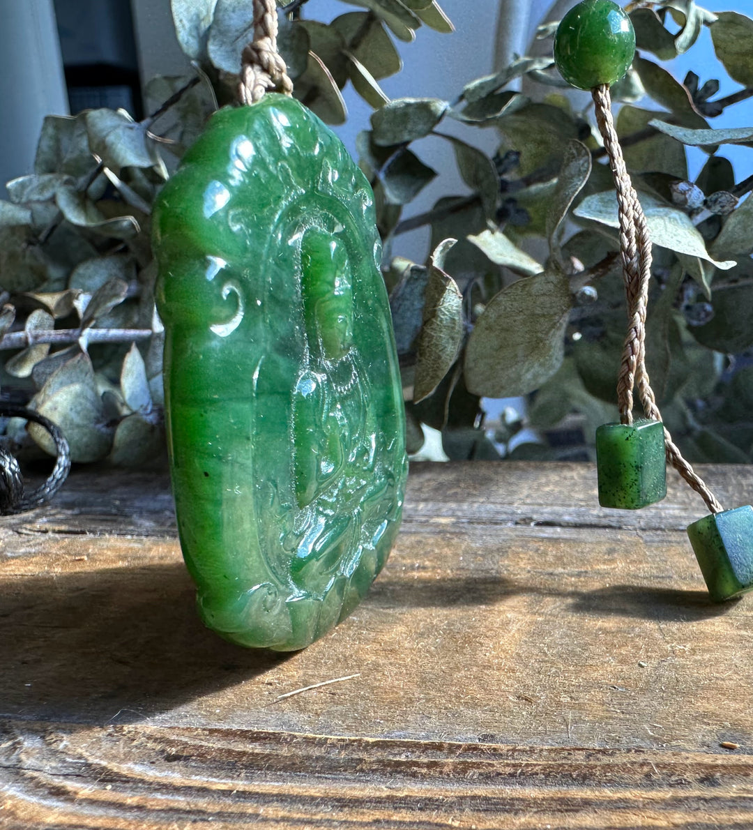 Green jade Guanyin pendant with braided cord and bead charms resting on wood with greenery in the background