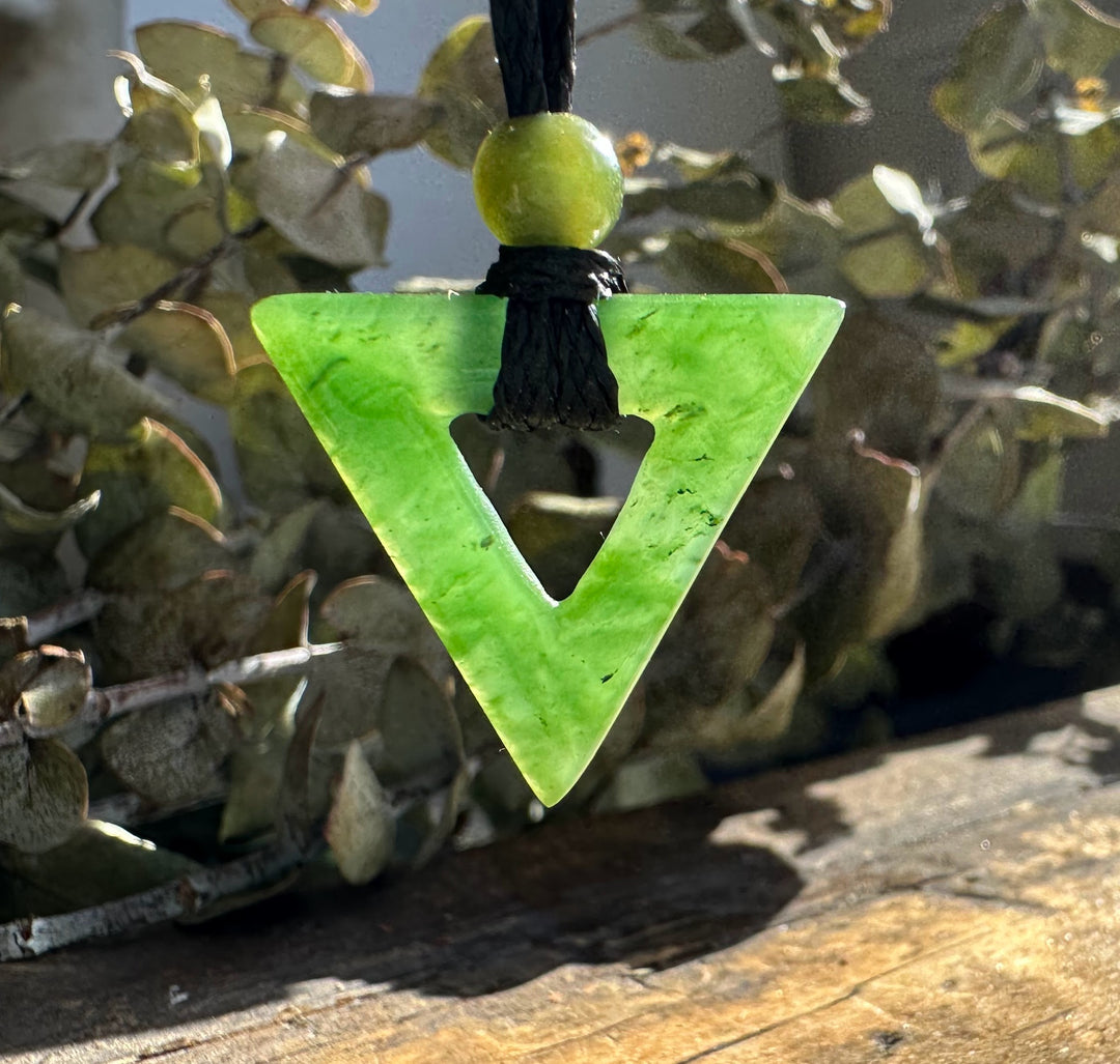 Close up of green jade triangle pendant with bead and black cord hanging among plants