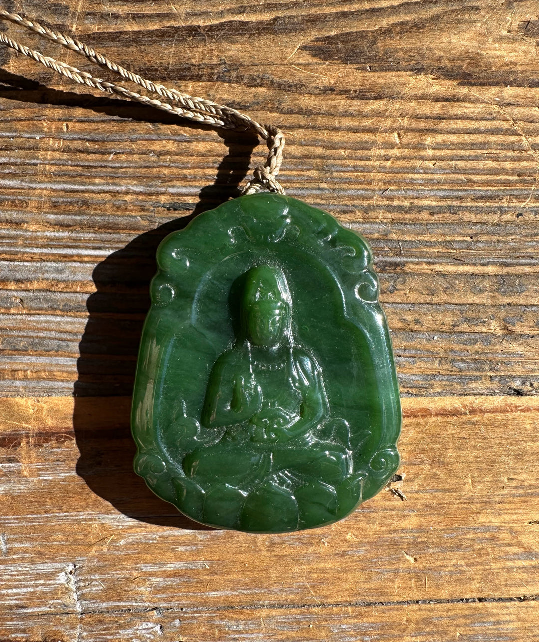 Close up of green jade Guanyin pendant showing the carved seated figure against a wooden surface.