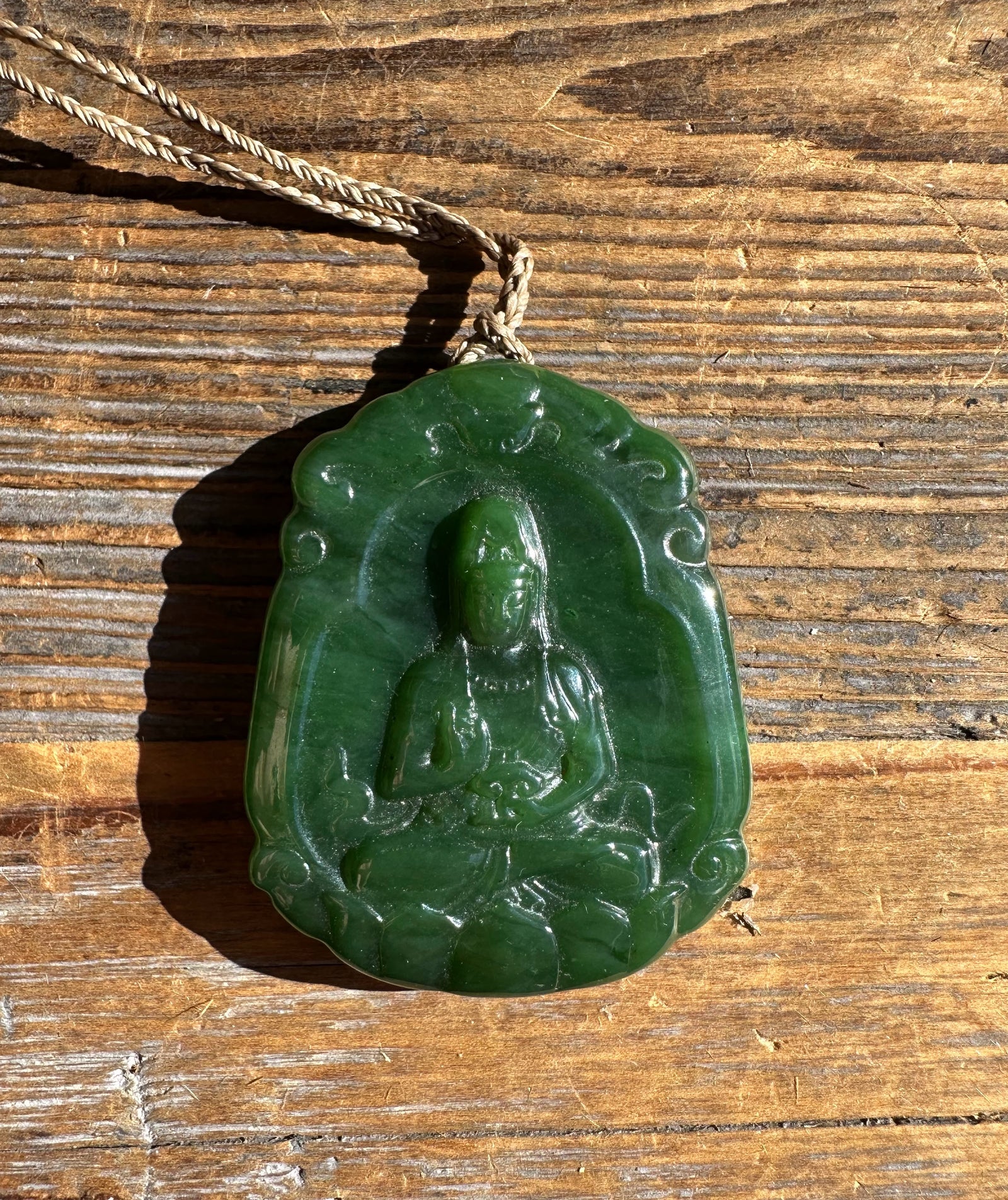 Close up of green jade Guanyin pendant showing the carved seated figure against a wooden surface.